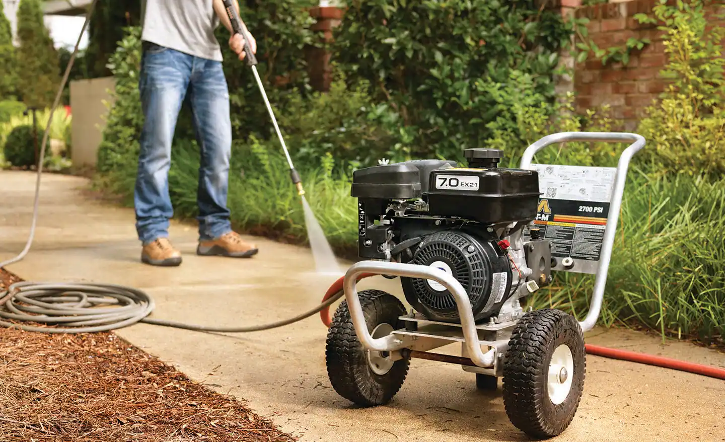 A homeowner kneeling on a concrete driveway, checking a gas pressure washer with a pull-start handle beside it, daylight real photo