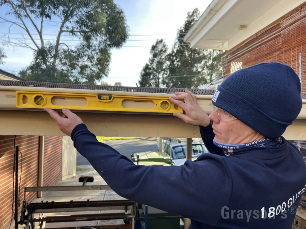 A homeowner on an extension ladder holding a small bubble level against the front lip of an aluminum gutter to check slope on a clear day