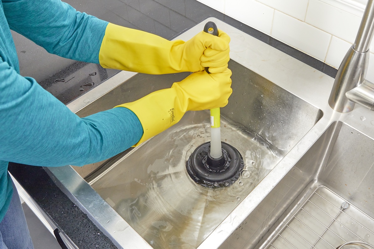 A homeowner plunging a stainless steel kitchen sink with water in the basin