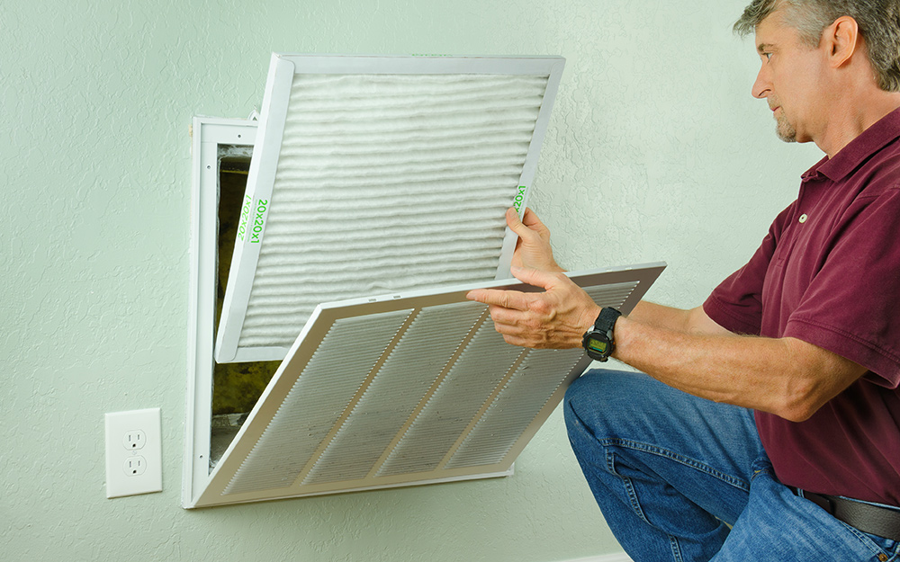 A homeowner sliding a pleated HVAC air filter out of a return grille in a residential hallway, realistic indoor lighting