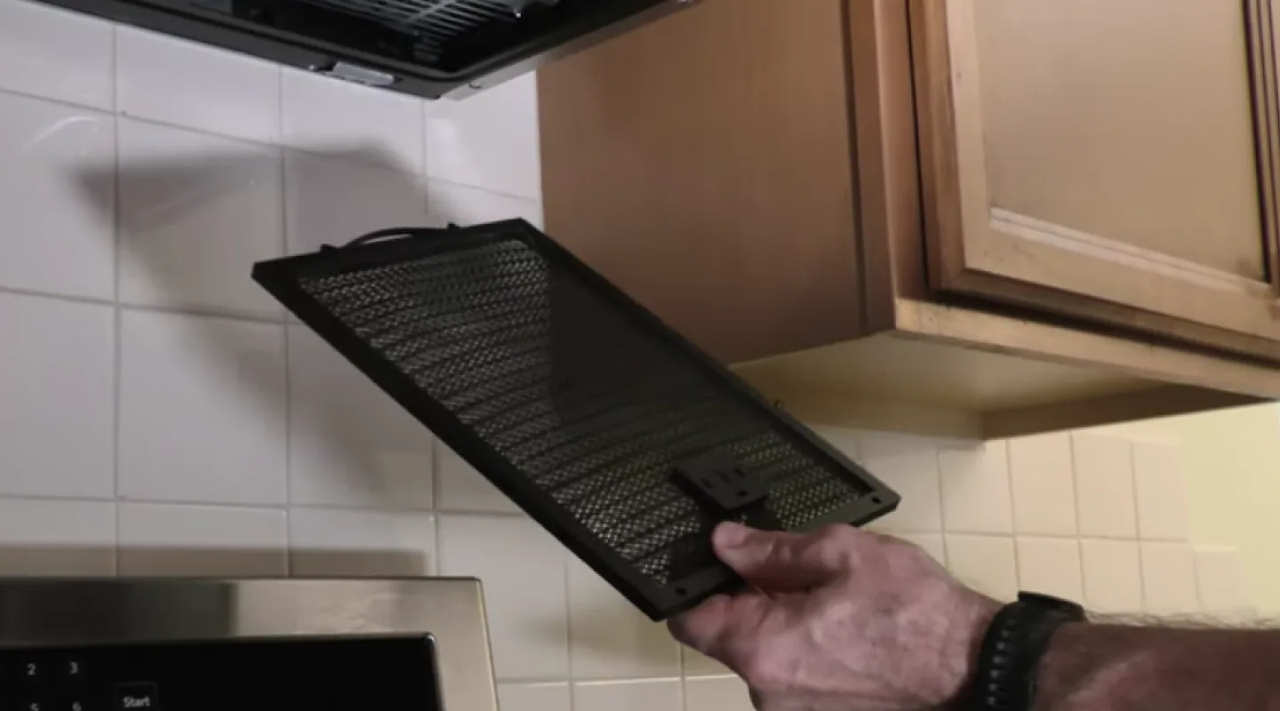 A homeowner standing at a kitchen range removing the metal grease filter from an over-the-range microwave