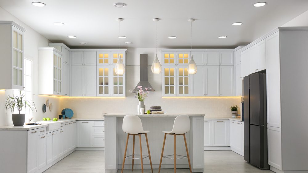 A homeowner standing in a kitchen looking up at a ceiling light while a refrigerator runs in the background