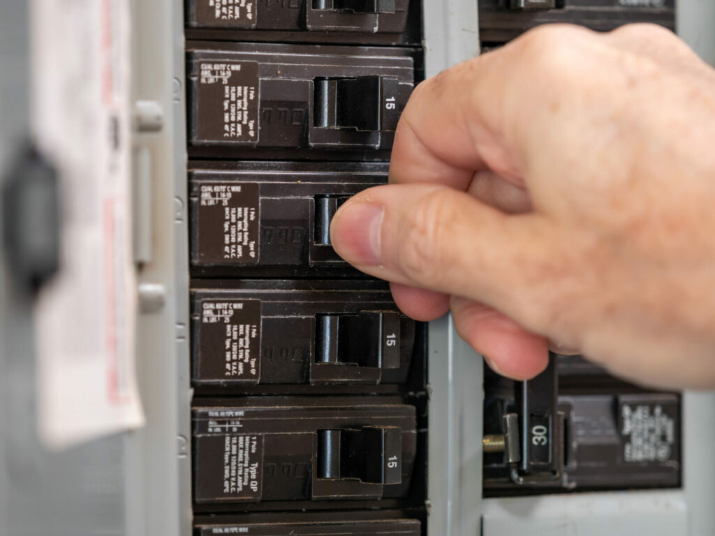 A homeowner standing in front of an open residential electrical panel in a kitchen-adjacent utility area, with one breaker in the tripped middle position, natural indoor light, realistic photo