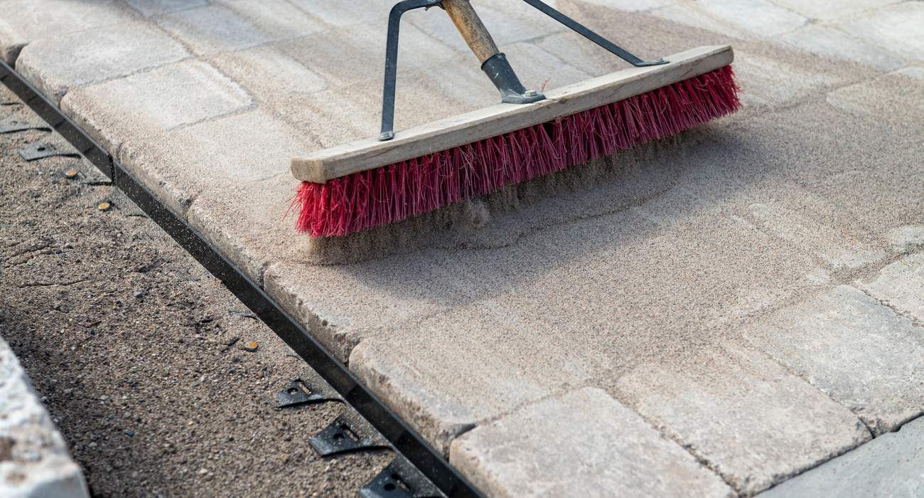 A homeowner sweeping tan polymeric sand across gray concrete patio pavers, with sand filling the joints on a sunny day, realistic photo