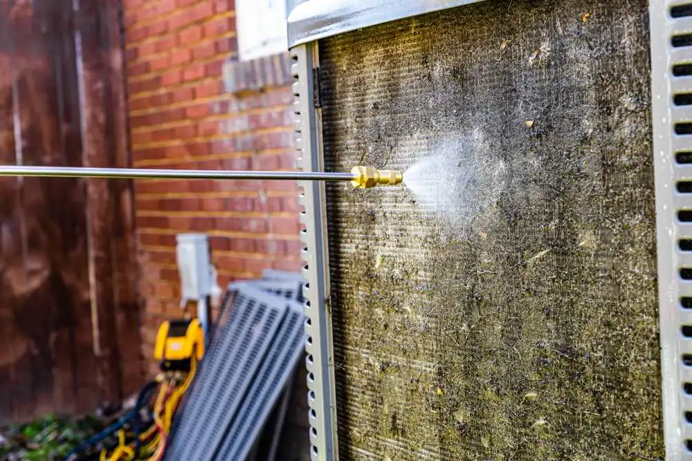 A homeowner using a garden hose to gently rinse the outdoor heat pump coil on a warm summer afternoon, real photography