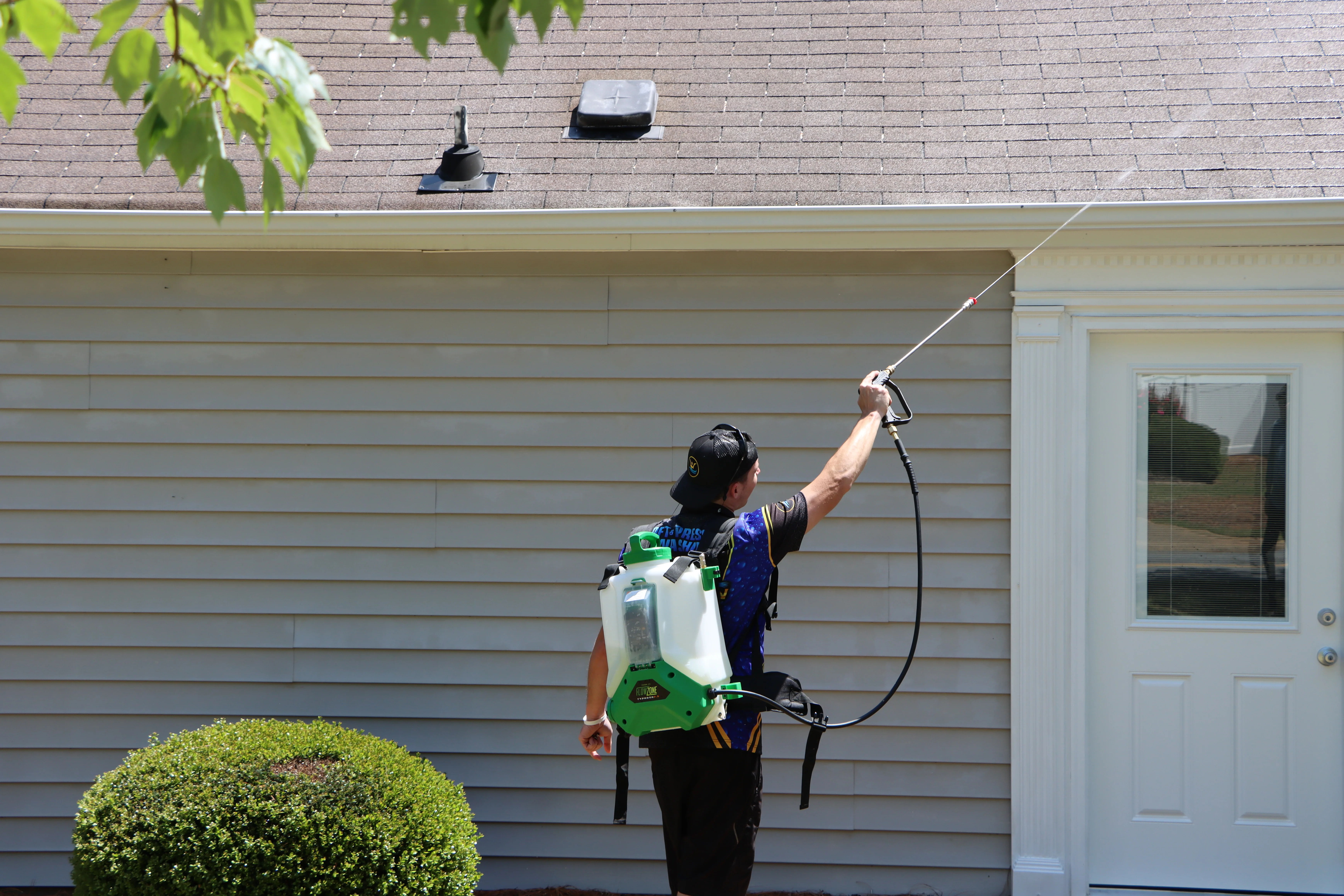 A homeowner using a hand pump sprayer to apply a gentle cleaning solution to vinyl siding on a sunny day, with a garden hose ready for rinsing
