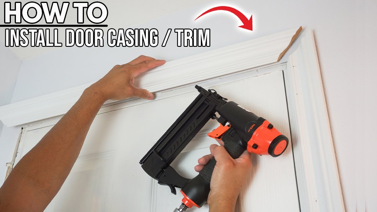 A homeowner using a pneumatic 16-gauge finish nailer to attach primed door casing to a wood jamb, workshop lighting, realistic photo