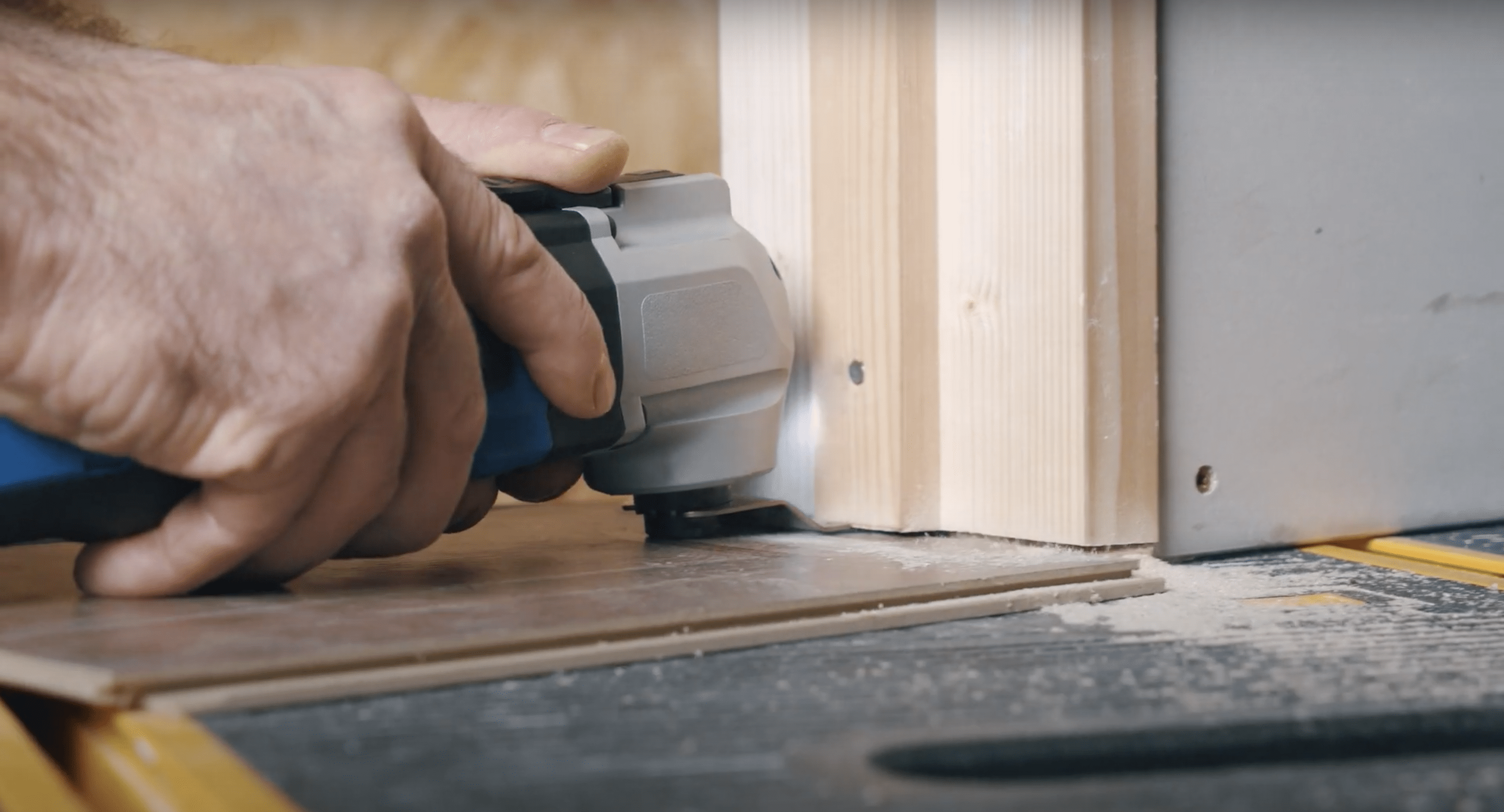 A homeowner using an oscillating multi-tool to undercut a door jamb using a scrap plank as a height guide