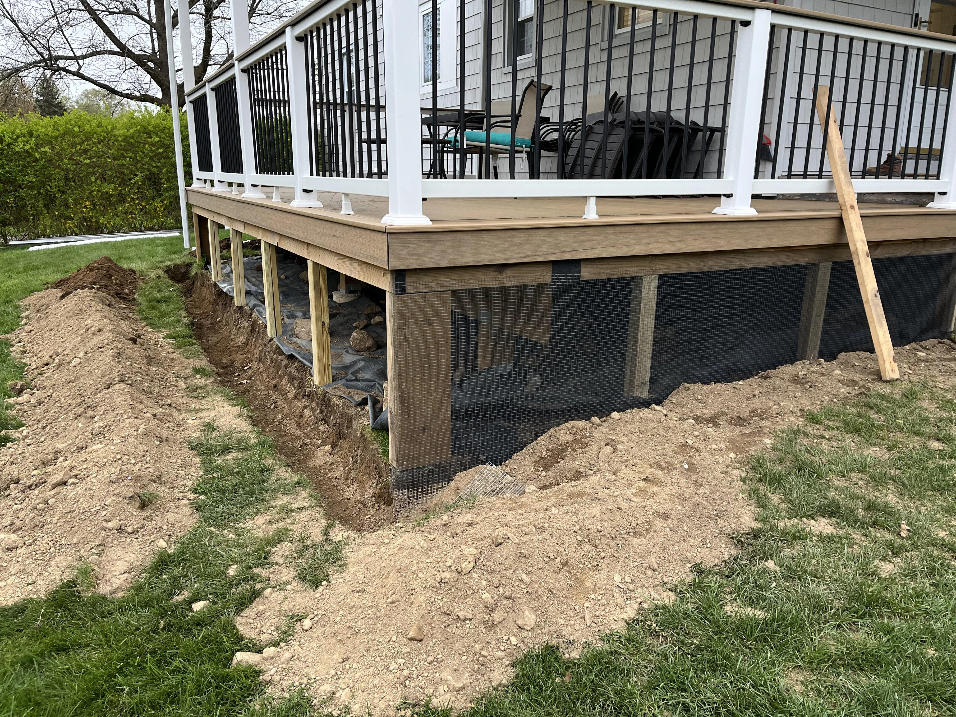 A homeowner wearing work gloves stapling galvanized hardware cloth along the bottom edge of a wooden deck to block animal entry