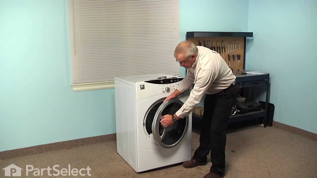 A household dryer with the door slightly open, showing the latch area and door strike in a real laundry room setting