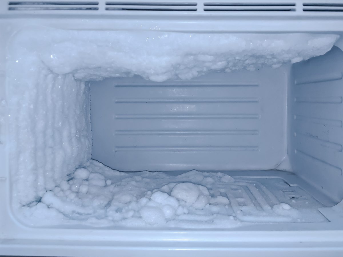 A kitchen freezer interior with heavy frost coating the back wall and ice buildup on a shelf edge, shot like a real home maintenance photo with natural indoor lighting