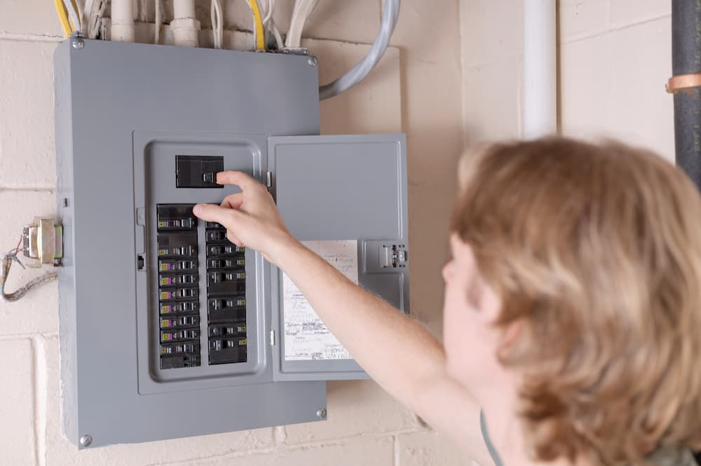 A licensed electrician kneeling in a kitchen using a handheld electrical tester at a countertop outlet while an open breaker panel is visible nearby, realistic indoor photo