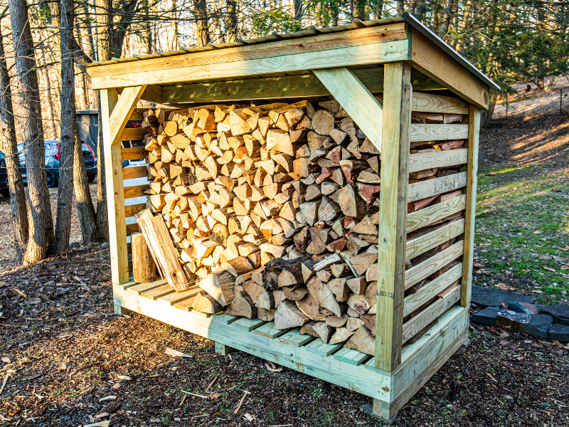 A neatly stacked firewood pile on a raised rack in a backyard, positioned away from a house, photographed in clear daylight
