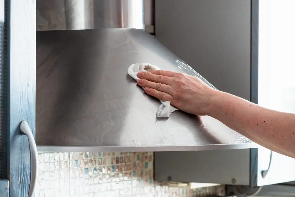 A pair of hands washing a white metal air vent cover in a stainless kitchen sink with soapy water, realistic home photo