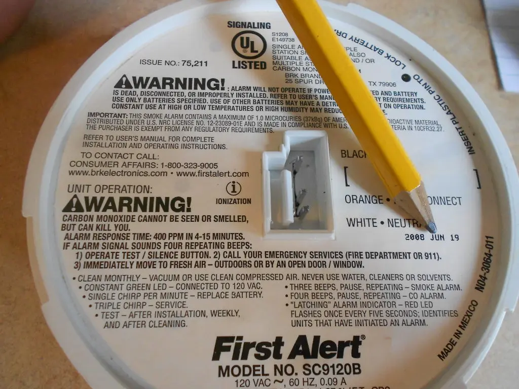 A person holding a carbon monoxide detector showing the back label with manufacturing date and replacement information in a home environment