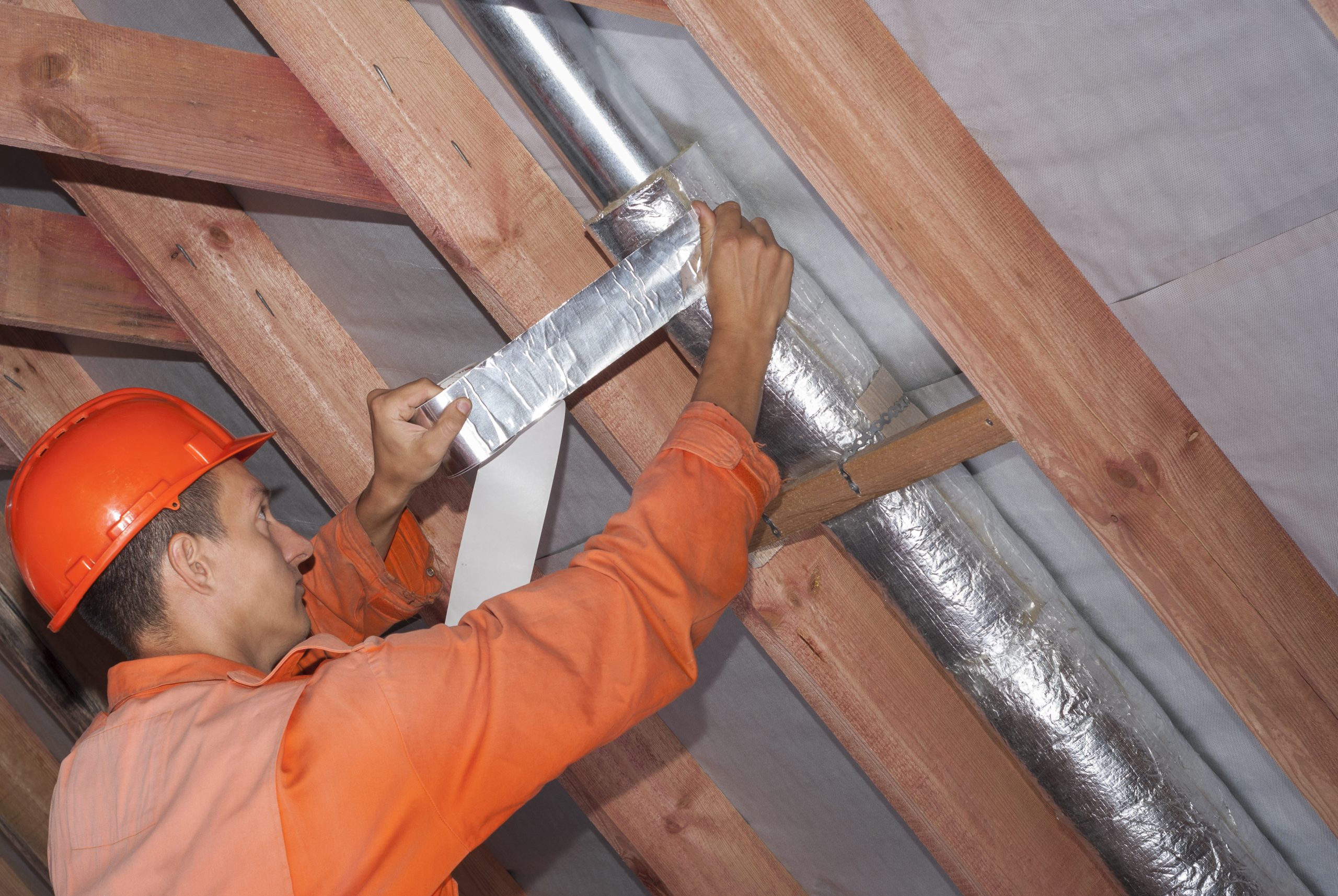 A person in an attic applying foil HVAC tape to a metal duct joint near insulation, realistic home maintenance photo