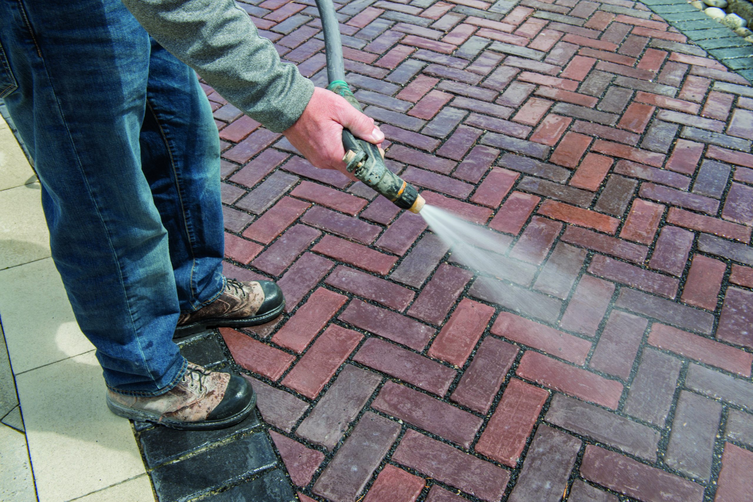 A person pressure washing a patio, holding the wand at an angle to clean between concrete pavers with water spray visible, realistic photo