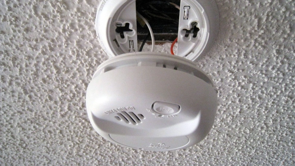 A person using a can of compressed air to blow dust out of the vent slots of a ceiling smoke alarm