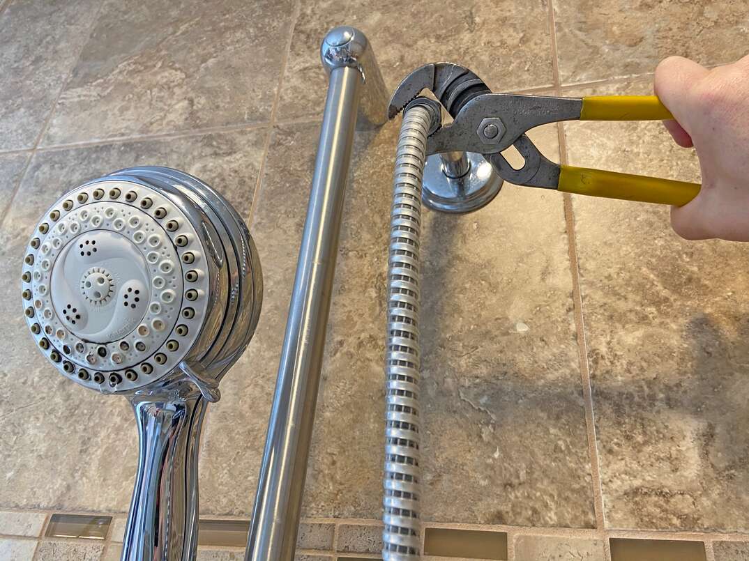 A person using a strap wrench to loosen a chrome shower arm at the wall in a tiled shower, close-up real photo