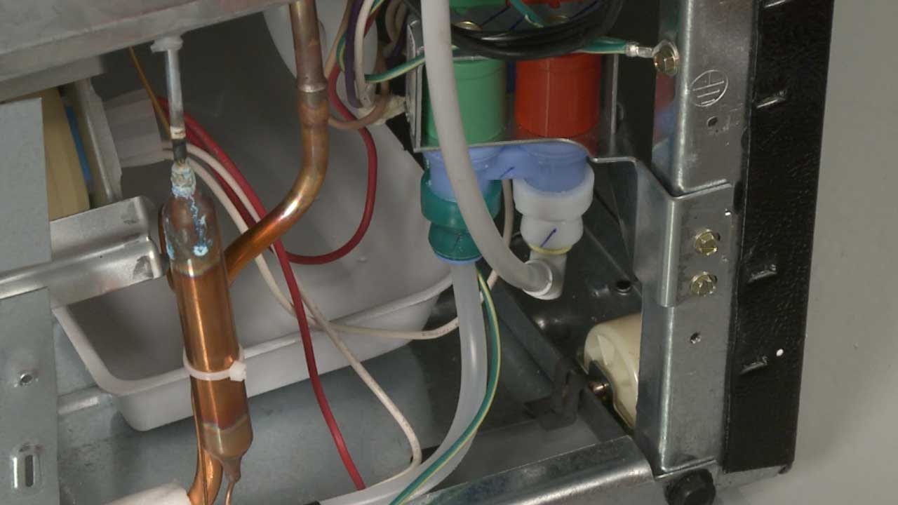 A photo of the back lower portion of a refrigerator showing a water inlet valve with two water line connections and electrical connectors, workshop lighting