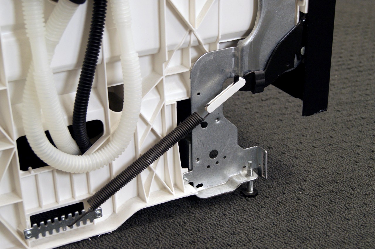 A photo of the lower side area of a built-in dishwasher with the toe-kick removed, showing the door hinge arm and spring connected to the frame