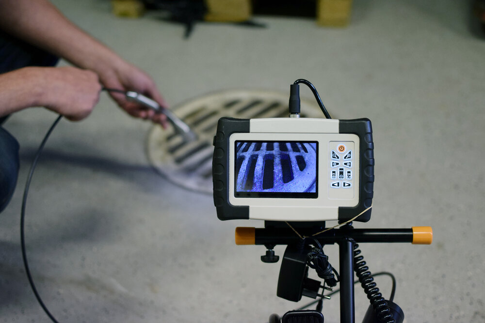 A plumber feeding a sewer camera cable from a reel into an outdoor cleanout while watching a monitor on the ground, realistic photo