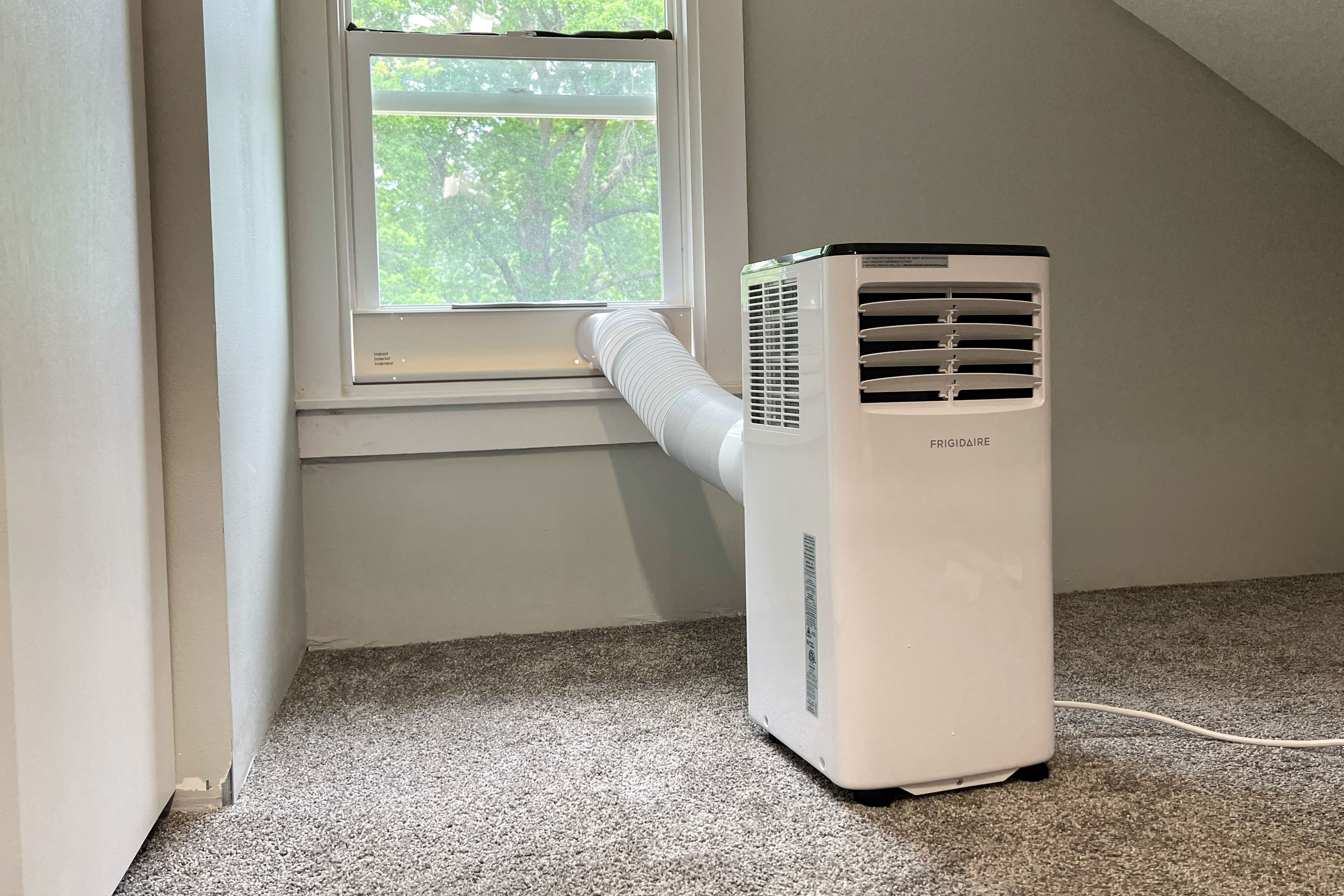 A portable air conditioner unit on a hardwood floor next to a window with an exhaust hose connected, photographed in a real home