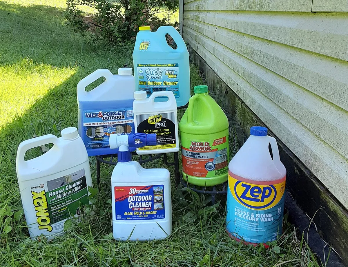 A pump sprayer, oxygen bleach container, soft bristle brush, garden hose, and nitrile gloves laid out on a driveway next to a house