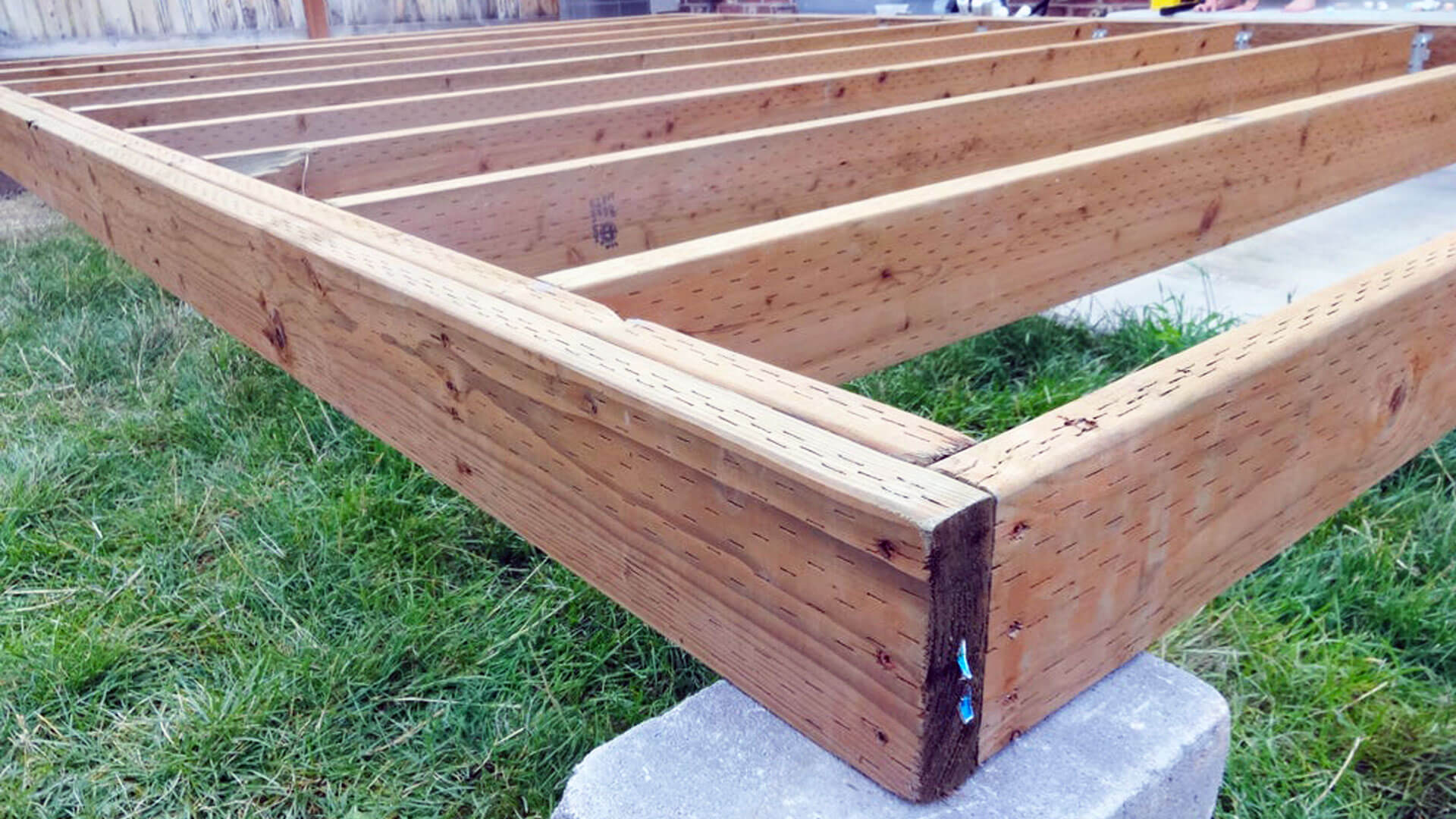 A raised backyard deck under construction with exposed pressure-treated joists, a double beam on posts, and a ledger attached to the house