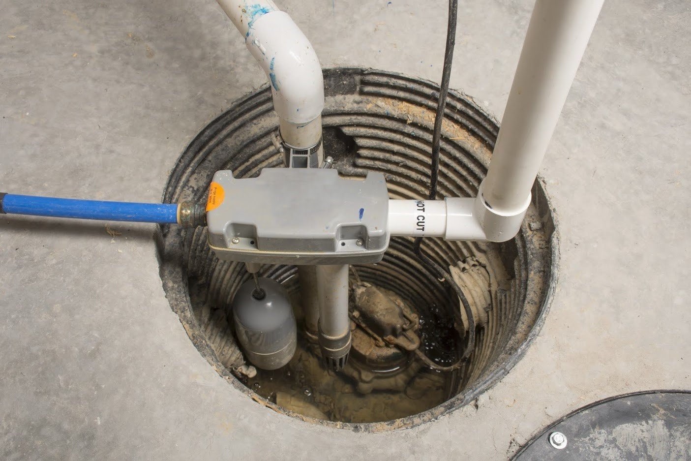 A real basement bathroom sewage ejector pump pit with the lid removed, showing the pump basin and plumbing connections in a concrete floor, indoor home photo
