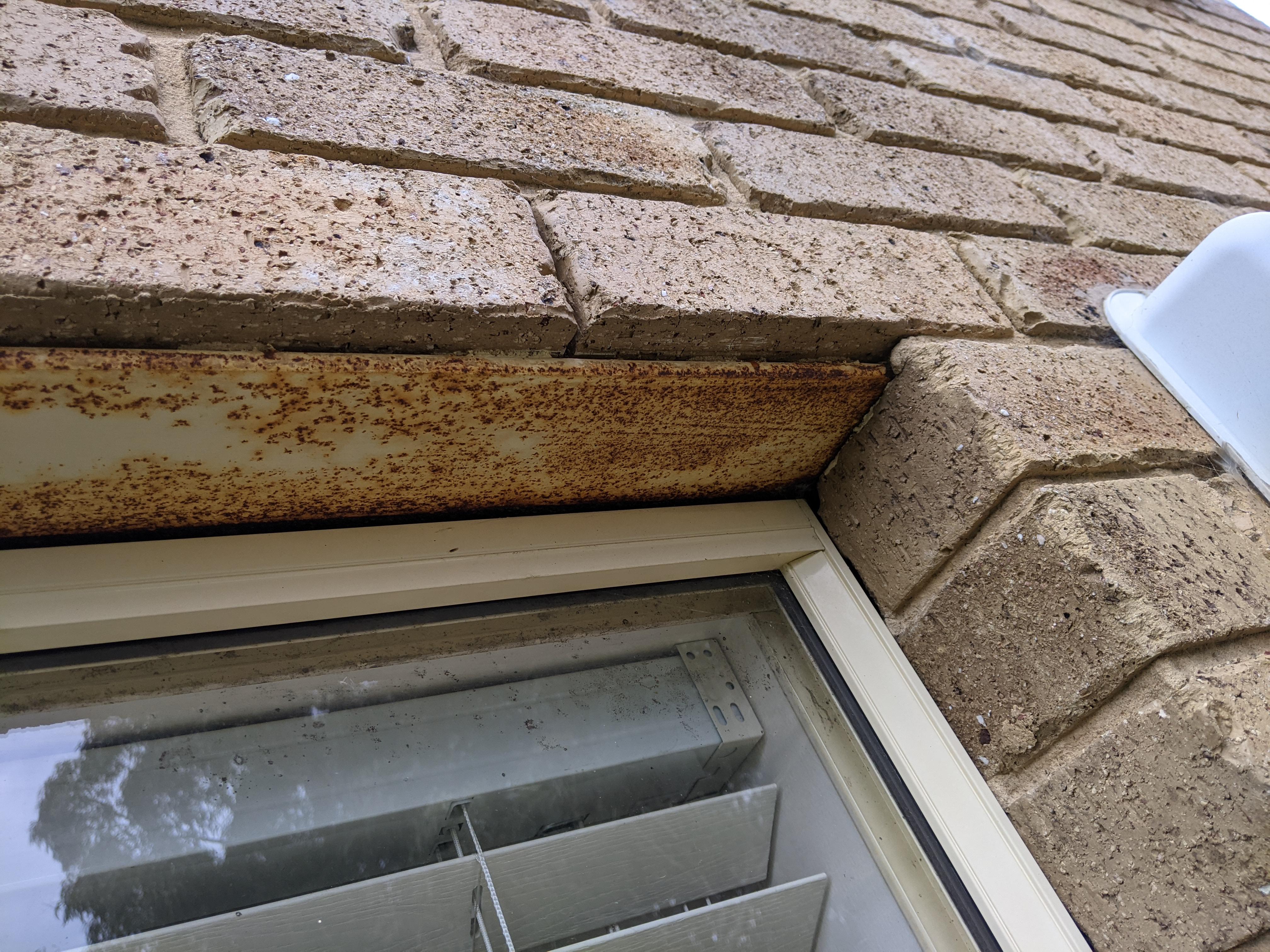 A real close-up photo of a brick wall above a window showing rust staining on the steel lintel and cracked mortar joints nearby