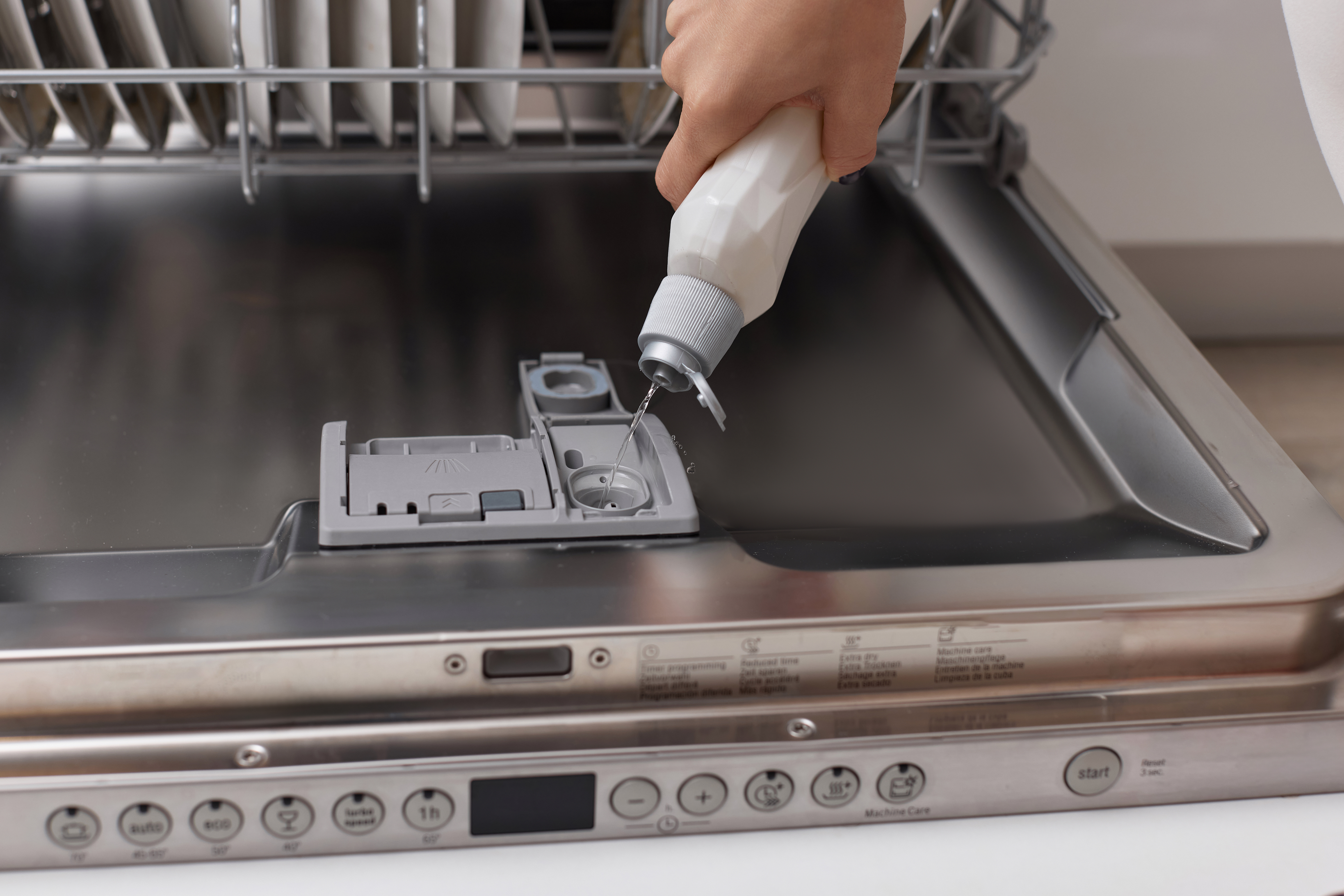 A real close-up photo of an open dishwasher door showing the rinse aid dispenser cap area on the inner door, with visible water droplets and typical kitchen lighting