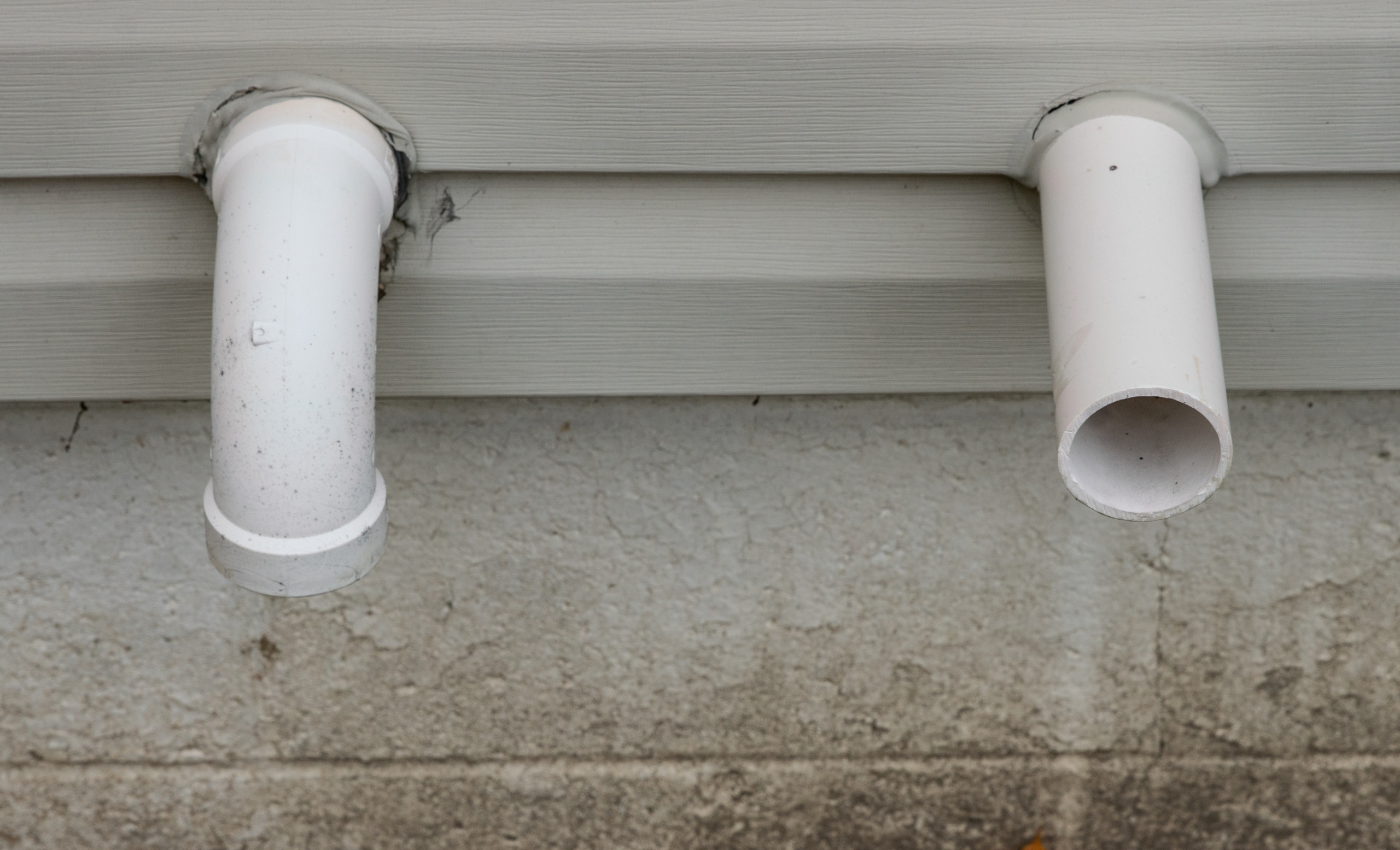 A real exterior photo of two white plastic pipes exiting a house side wall with standard vent elbows