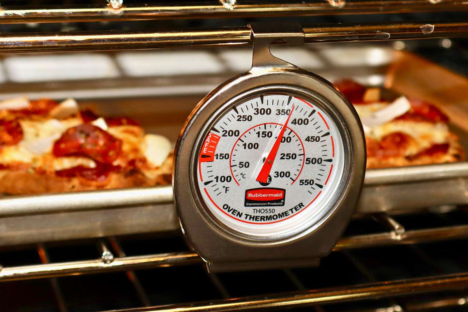 A real home kitchen oven with the door open and a standalone analog oven thermometer sitting on the center rack, photographed in natural indoor light