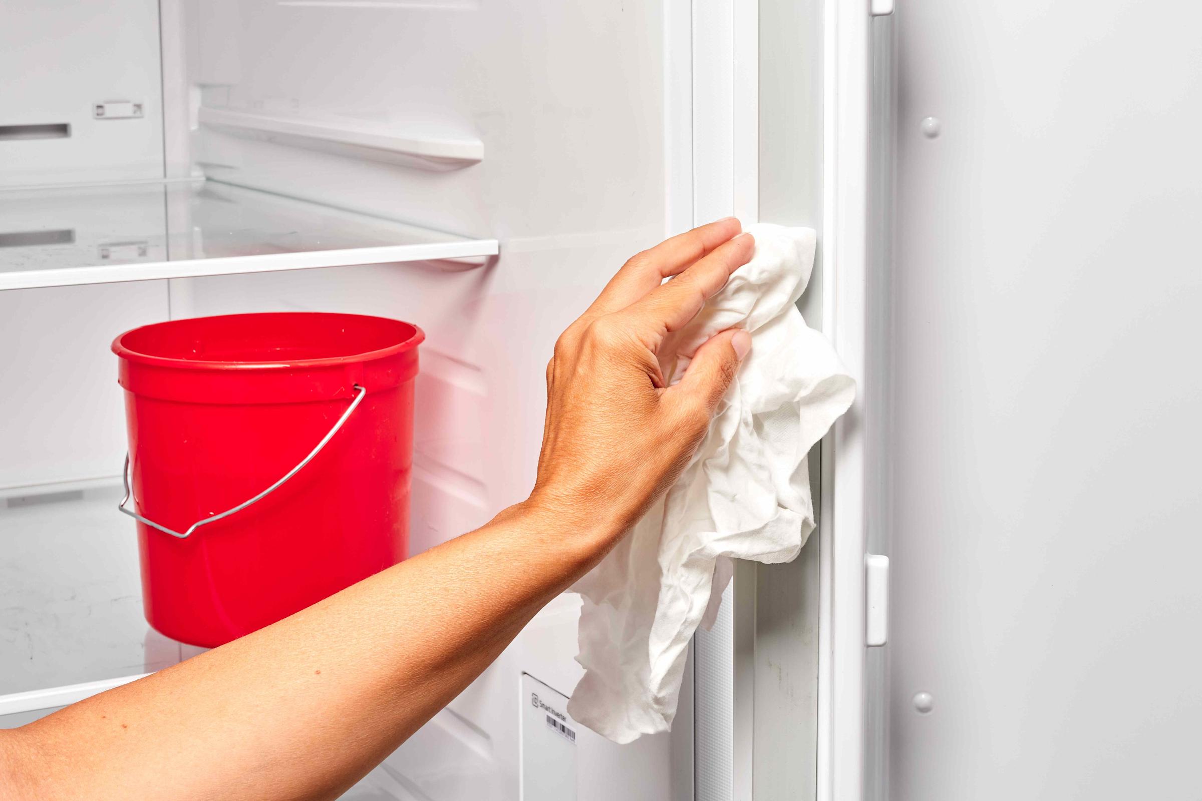 A real kitchen freezer door slightly open while a person wipes the rubber door gasket with a cloth and warm soapy water, close-up photo