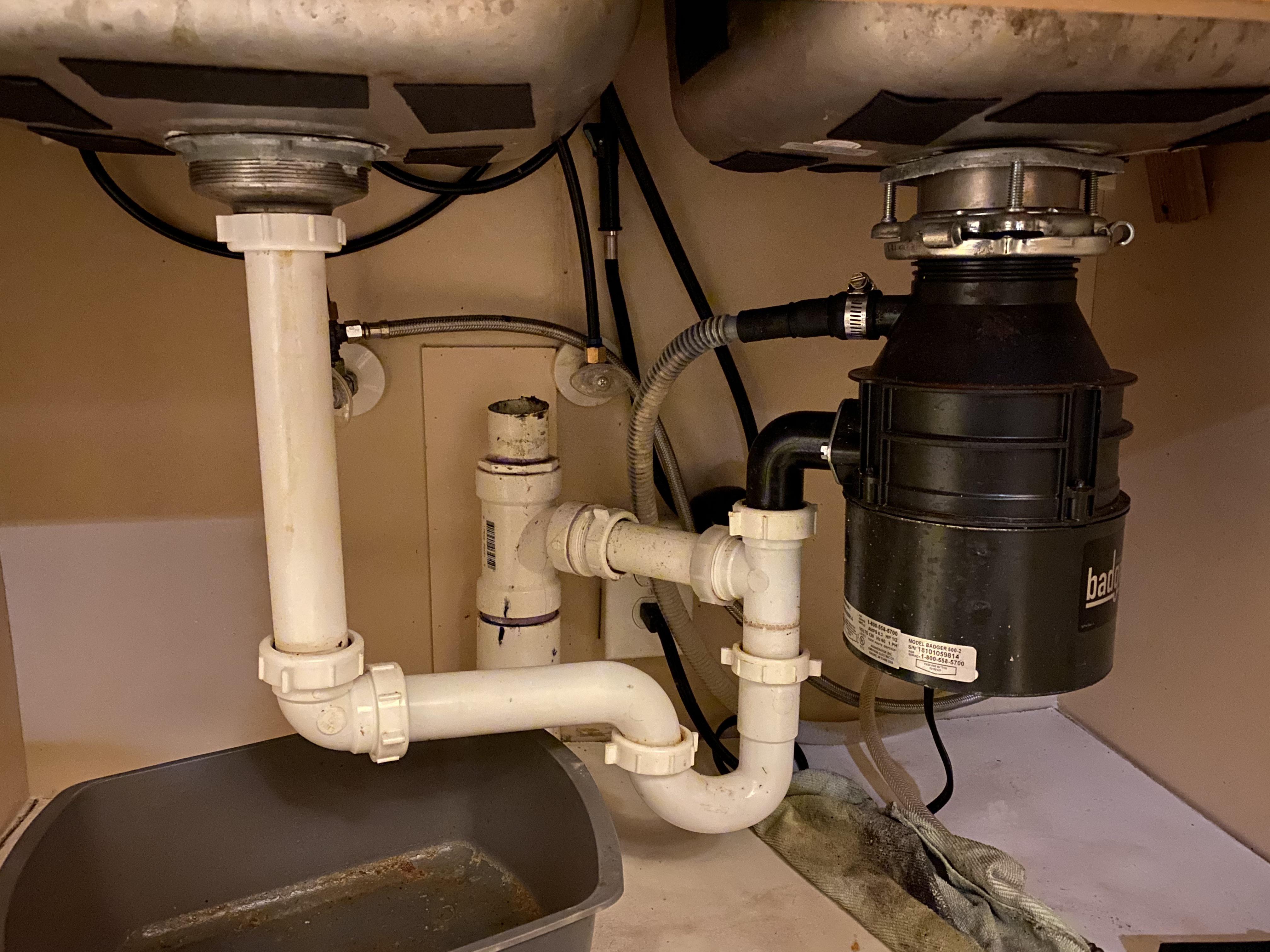 A real kitchen sink cabinet interior with a garbage disposal, drain pipes, and a bucket placed underneath, photographed during a home repair