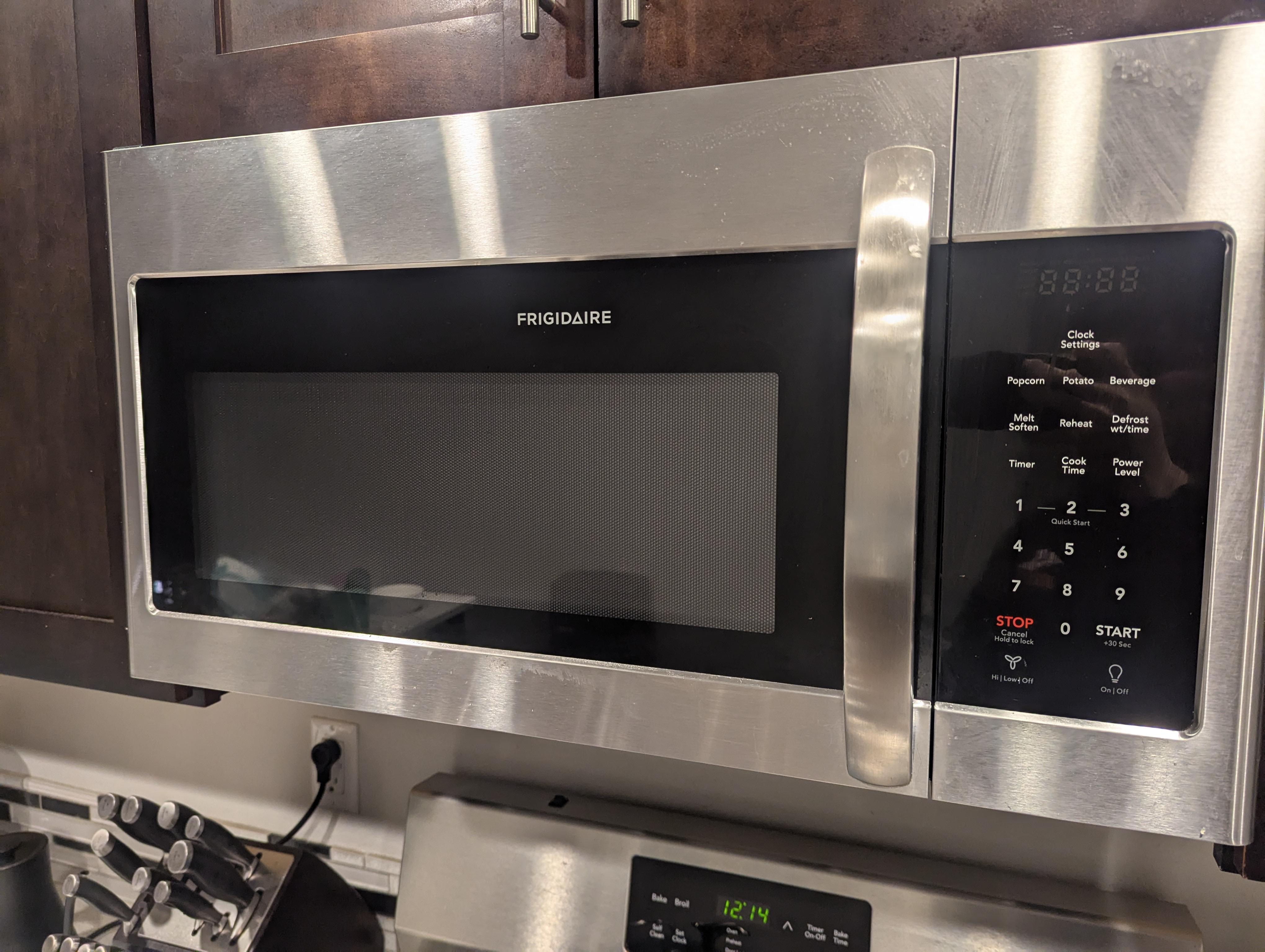 A real kitchen with an over-the-range microwave above a stove, the microwave display off as if it lost power, with a nearby electrical panel door open in the background, natural indoor lighting