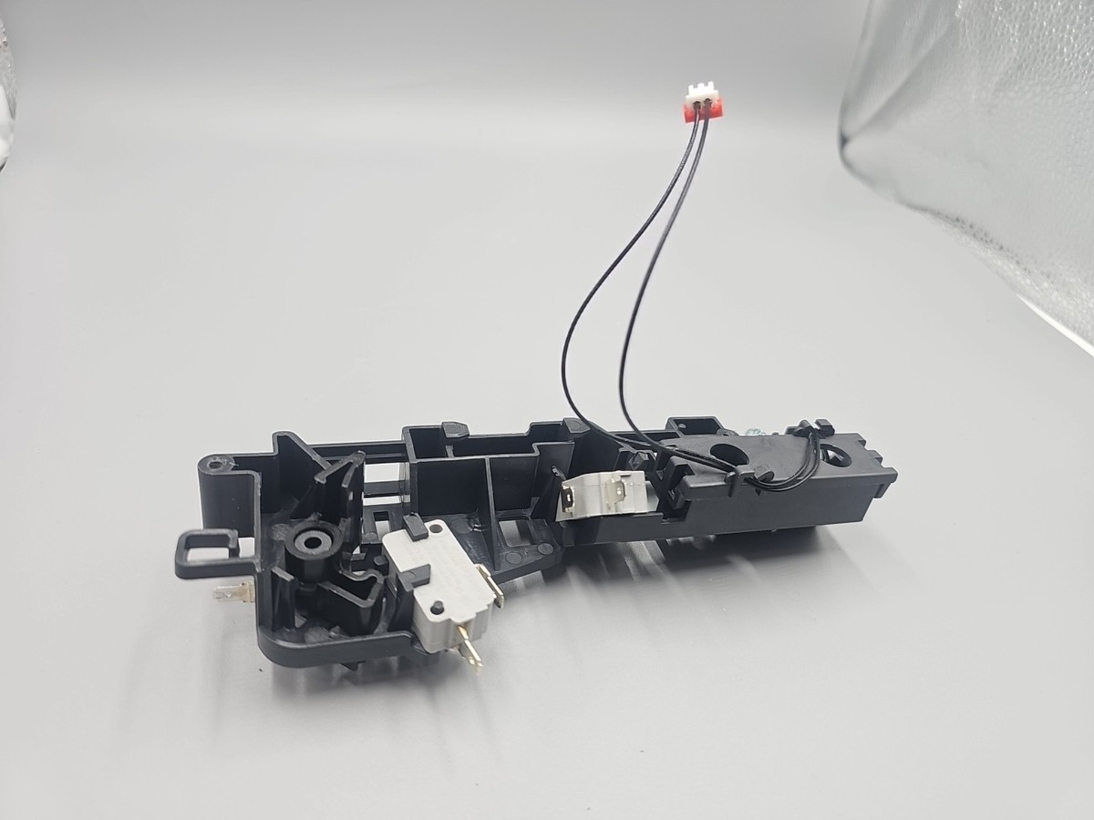 A real-life photo of a hand using a small toothbrush to clean the latch receiver area on a microwave frame in a home kitchen