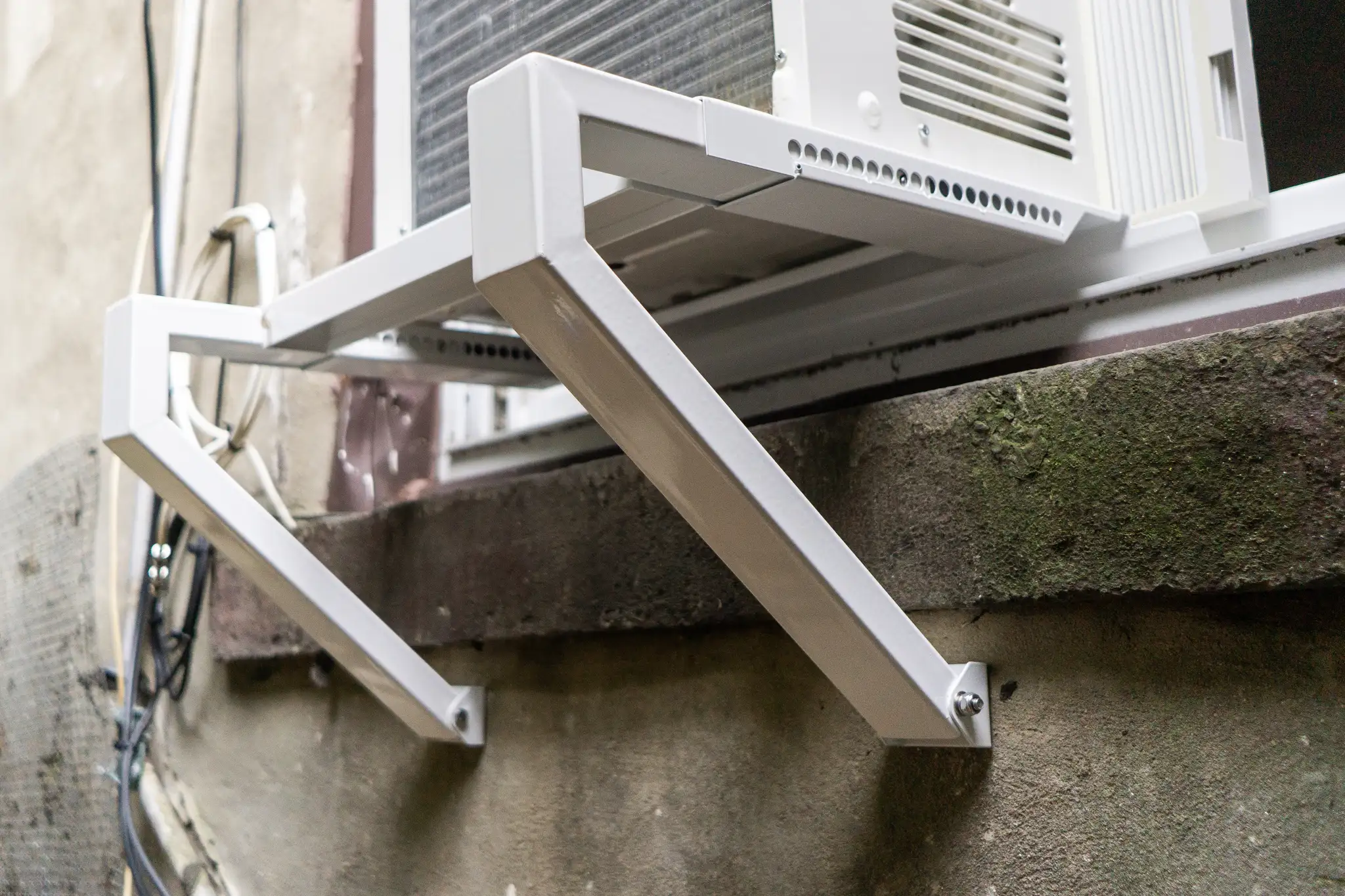 A real-life photo of the exterior side of a window air conditioner resting on a metal support bracket attached below the window, showing the unit supported securely with the recommended drainage angle