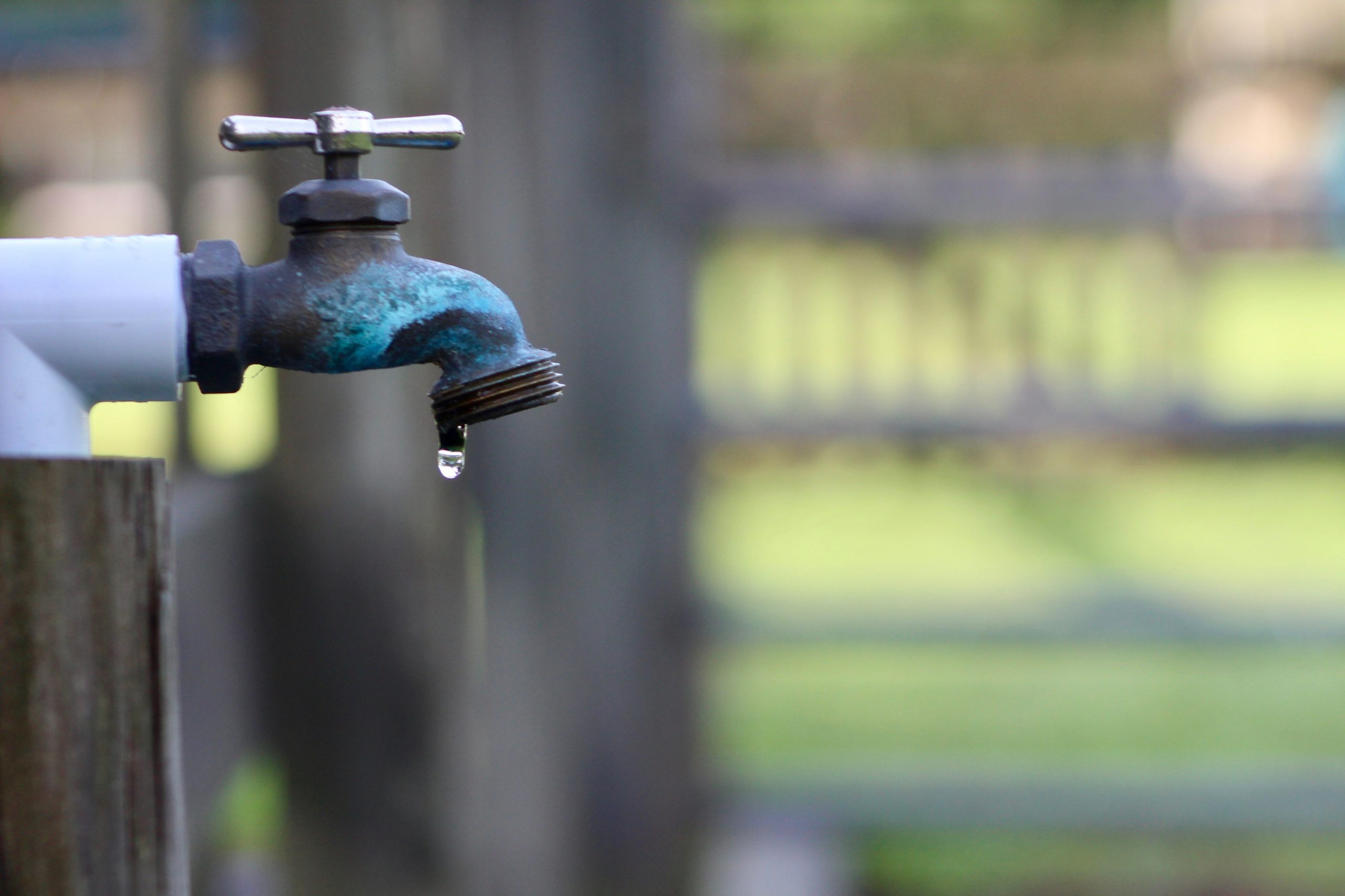 A real outdoor hose bib on a brick wall with water streaming from the spout into a puddle on a concrete patio in daylight