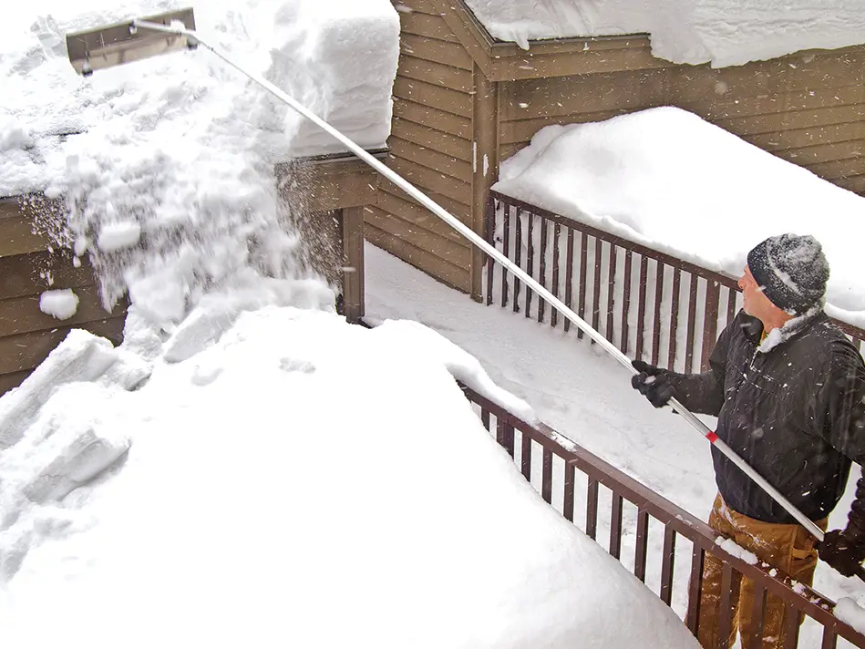 A real outdoor photo of a homeowner standing on the ground using a long-handled roof rake to pull snow off the lower edge of a snowy roof