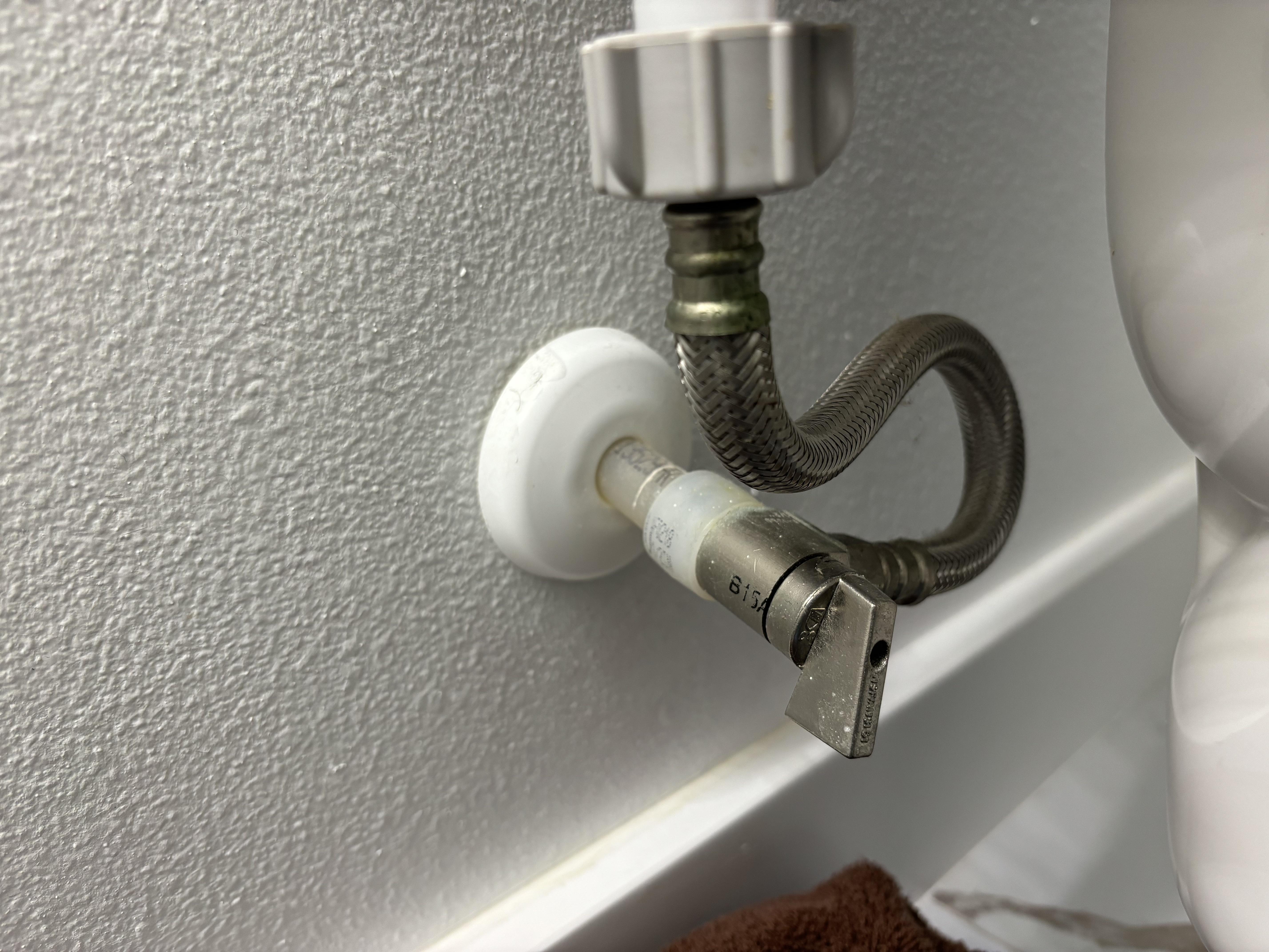 A real photo behind a toilet showing a braided water supply line bent sharply and kinked near the shutoff valve