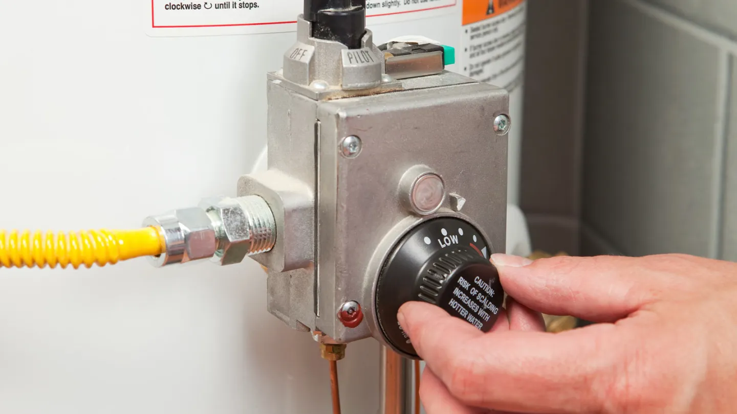 A real photo close-up of a gas water heater control valve with temperature dial and gas line connection in a home utility room, sharp focus on the control knob