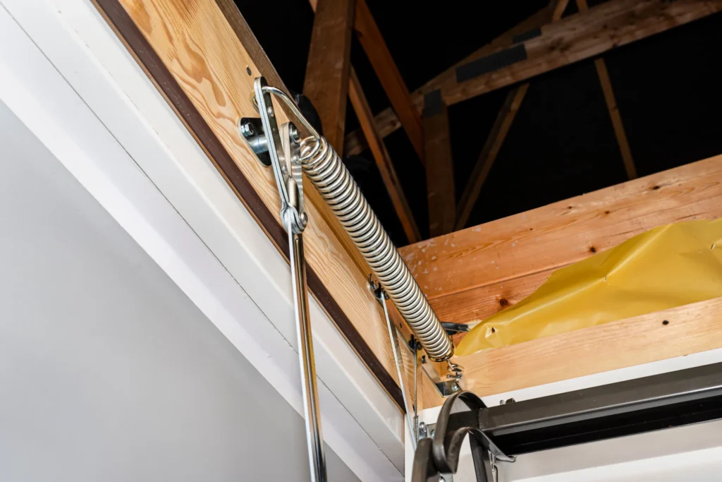 A real photo closeup of an extension spring on the side of a pull-down attic ladder, attached to a metal bracket and wood frame