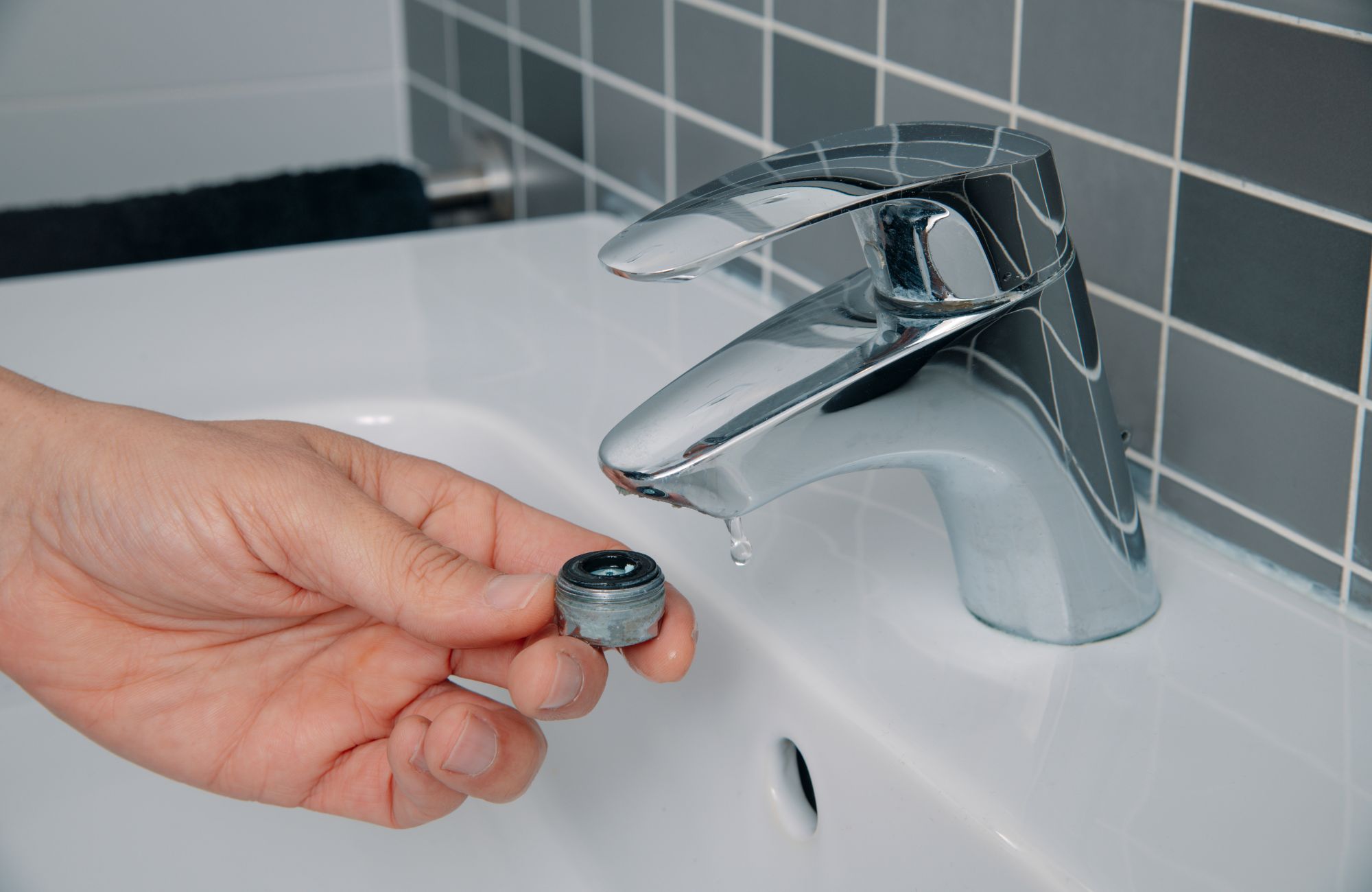 A real photo of a bathroom sink faucet aerator removed and held in a hand, showing trapped reddish-brown grit and mineral debris