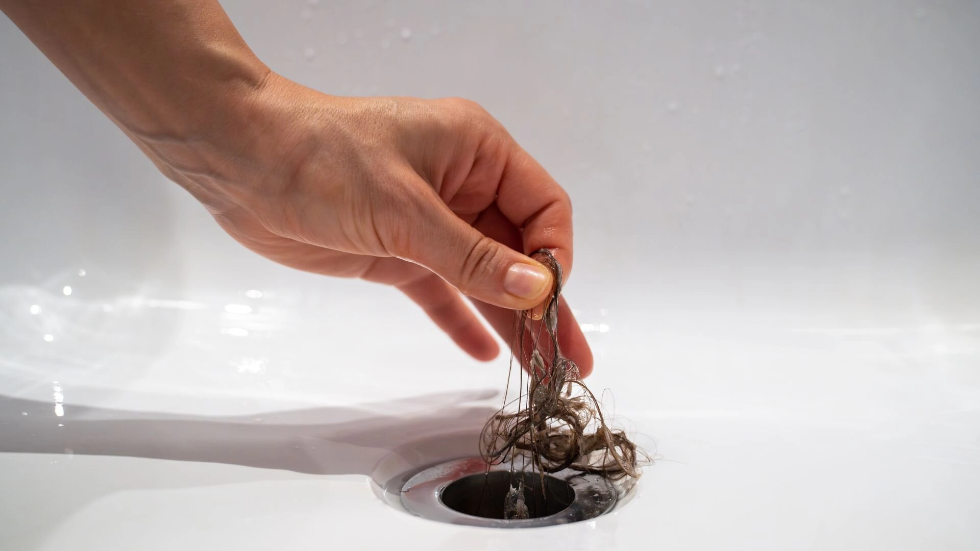 A real photo of a bathtub drain with the stopper removed, showing a clump of hair being pulled out with gloved hands and needle-nose pliers