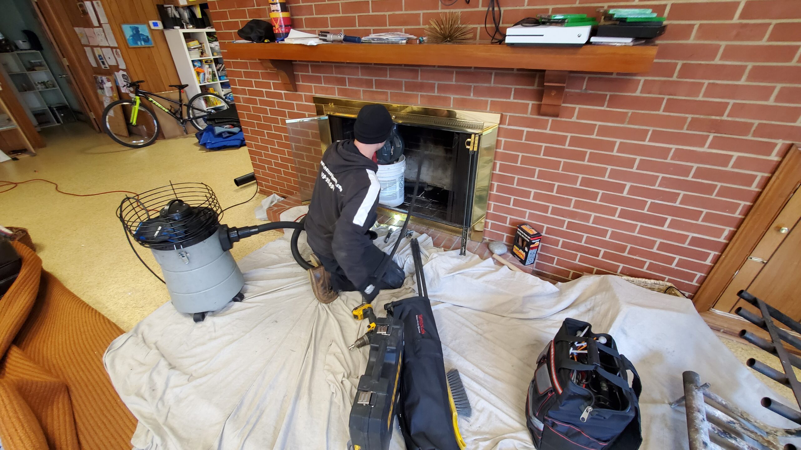 A real photo of a certified chimney sweep wearing protective gear using a chimney brush and rods near a fireplace opening inside a home