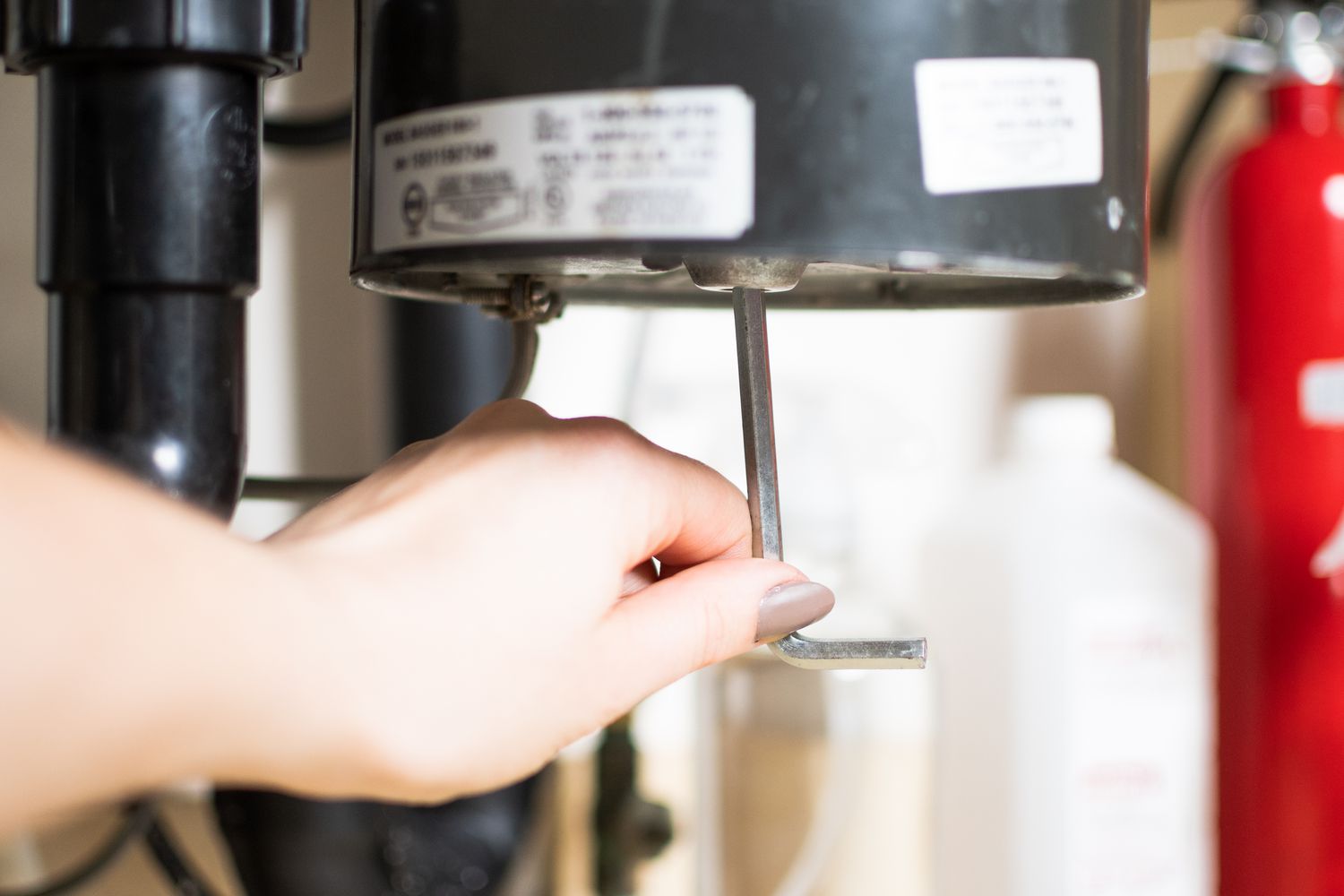 A real photo of a hand holding an L-shaped hex wrench next to the bottom of a garbage disposal under a kitchen sink, with the hex socket centered on the disposal base