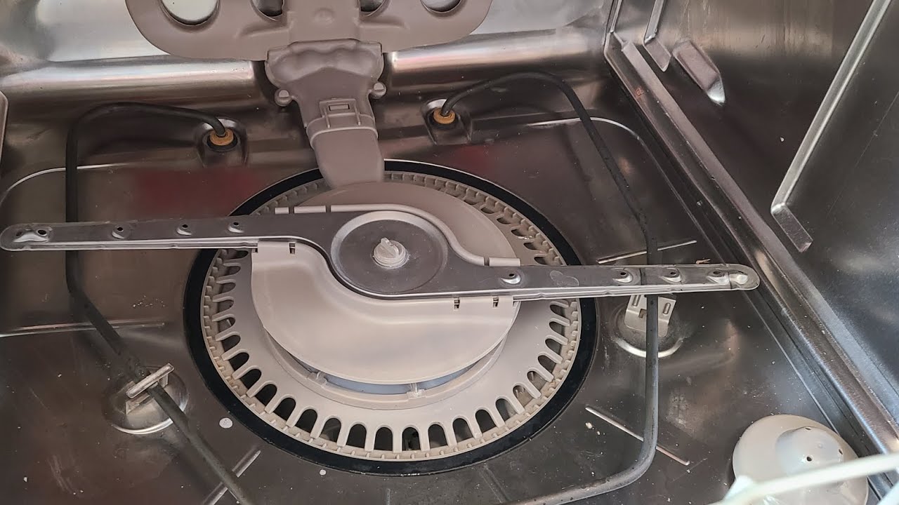 A real photo of a hand lifting a cylindrical dishwasher filter out of the bottom basin with the lower rack removed