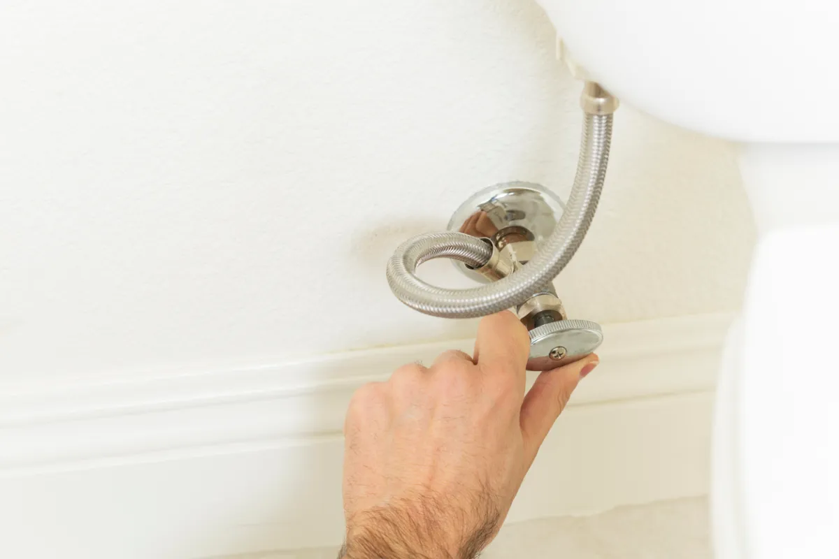 A real photo of a hand turning a toilet shutoff valve next to a bathroom wall, close-up, natural indoor lighting